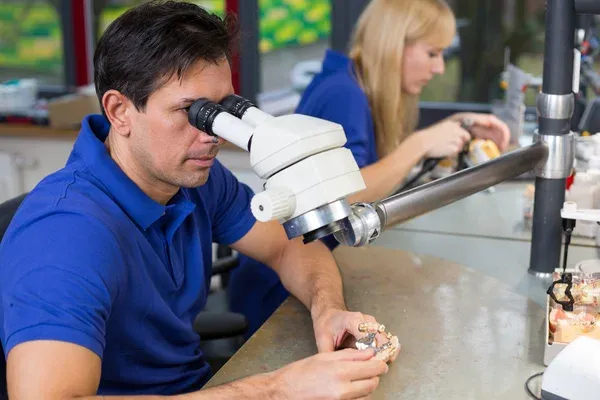 A dental technician examining a restoration under a stereo microscope at the bench.